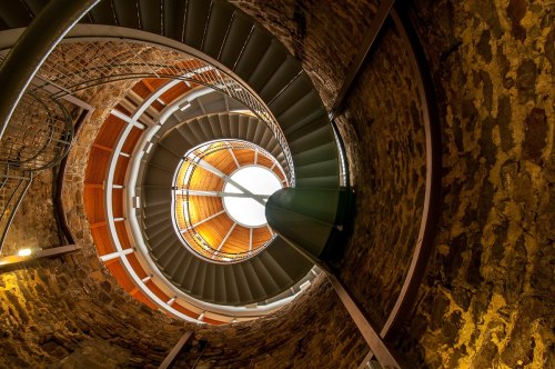 A spiral staircase viewed from below, featuring stone walls and a circular skylight at the top.