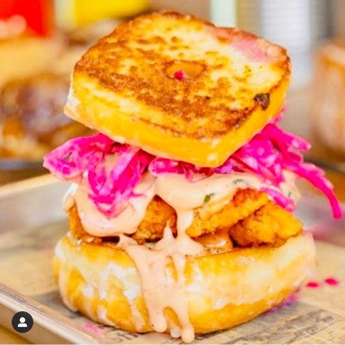 A stacked sandwich featuring fried chicken, pink slaw, and a glazed donut as the bun, drizzled with sauce.