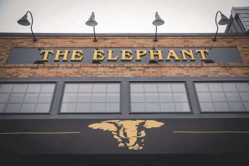 Sign for "The Elephant" featuring an elephant graphic, mounted on a brick building with windows above.