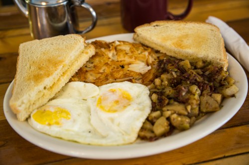 A plate of breakfast featuring two fried eggs, hash browns, diced potatoes with meat, and two slices of toast.