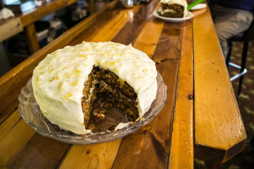 A sliced carrot cake with cream cheese frosting on a glass plate, set on a wooden table.