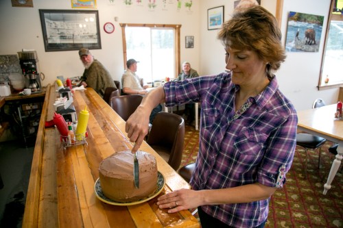 A woman in a plaid shirt slices a chocolate cake at a rustic diner, with patrons seated in the background.
