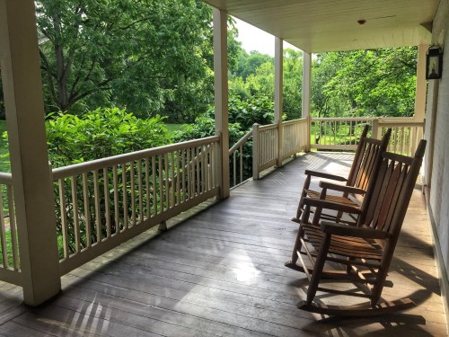 A peaceful porch with two rocking chairs overlooking a lush green garden. Sunlight filters through the trees.