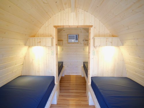 Cozy wooden interior of a small cabin with two beds, shelves, and an air conditioning unit. Warm lighting enhances the space.