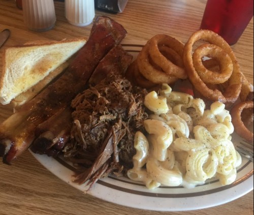 A plate of barbecue ribs, pulled pork, macaroni and cheese, onion rings, and toast on a wooden table.