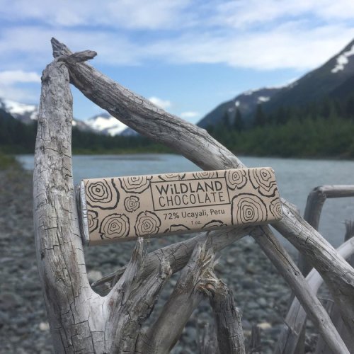 A chocolate bar from Wildland Chocolate rests on driftwood by a river, with mountains and a blue sky in the background.