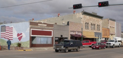 A small town street with colorful storefronts, a mural featuring a heart and American flag, and parked cars.