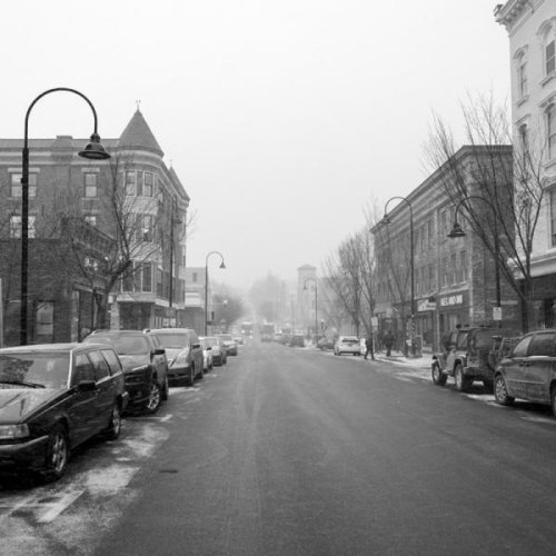 A snowy street scene with parked cars, historic buildings, and bare trees under a gray sky.