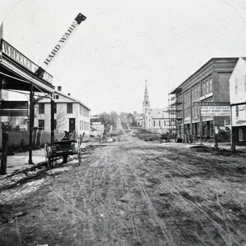 Historic black-and-white photo of a dirt street lined with buildings, a church steeple in the distance, and a horse-drawn cart.