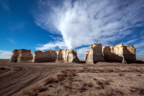 A vast landscape featuring towering rock formations under a blue sky with scattered clouds. Dry terrain in the foreground.