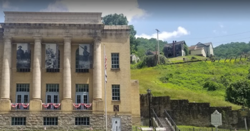 Historic building with columns, banners, and an American flag, set against a green hillside and old structures in the background.