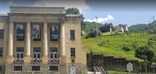 Historic building with columns, banners, and flags, set against a green hillside with old houses in the background.