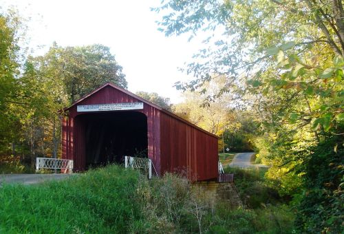 A red covered bridge surrounded by trees and grass, with a road visible in the background.