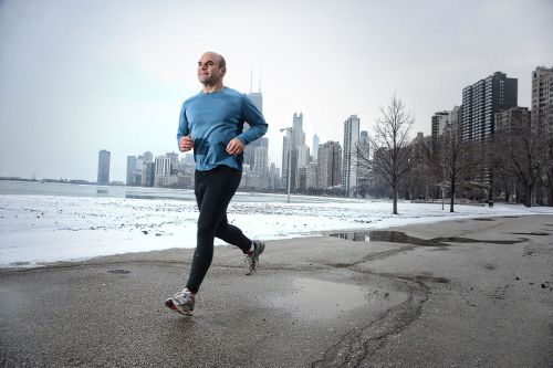 A man jogs along a snowy path with a city skyline in the background on a cloudy day.