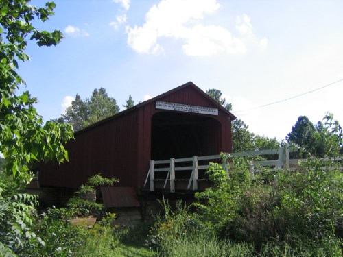 A red covered bridge surrounded by greenery under a blue sky with scattered clouds.