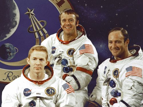 Three astronauts in space suits pose together, with a NASA logo and a starry background behind them.