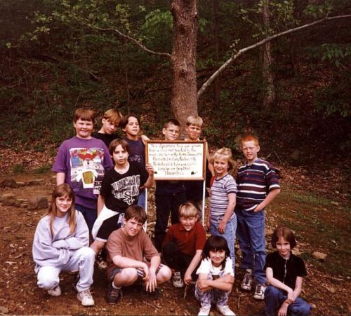 A group of children poses together outdoors, smiling in front of a sign on a tree.