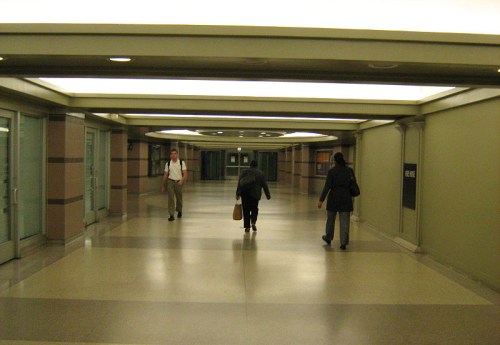 A spacious, well-lit corridor with three people walking, flanked by glass doors and smooth flooring.