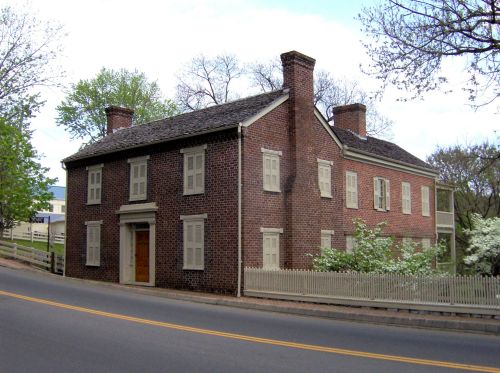 Historic brick house with white shutters, two stories, and a picket fence, set along a tree-lined road.