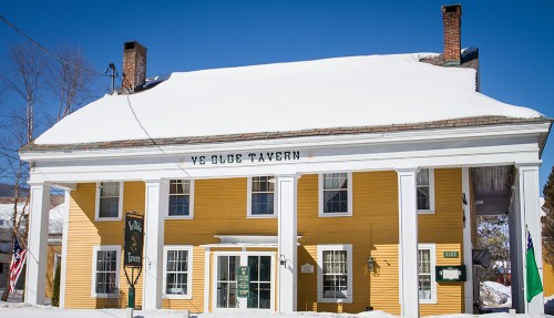A yellow tavern with white columns and a snow-covered roof, set against a clear blue sky.