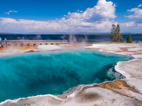 Vibrant blue hot spring surrounded by steam, with wooden walkways and a scenic lake under a partly cloudy sky.