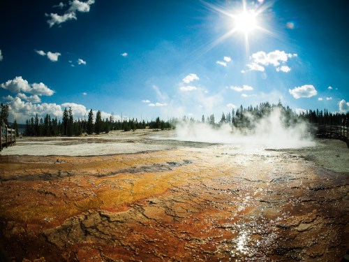 A vibrant landscape of Yellowstone with colorful mineral deposits, steam rising, and a bright sun against a blue sky.