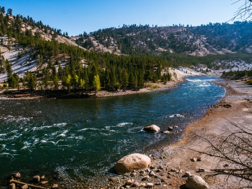 A serene river flows through a forested landscape, surrounded by mountains and rocky shores under a clear blue sky.
