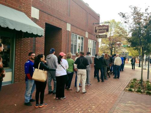 A long line of people waiting outside the Pancake Pantry, with a brick building and trees in the background.