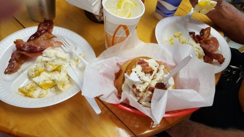 A table with two plates of scrambled eggs and bacon, and a sandwich topped with bacon in a basket, alongside a drink.