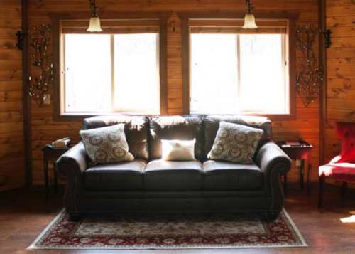 Cozy living room with a dark leather couch, decorative pillows, and wooden walls, illuminated by natural light.