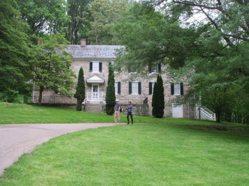 Two people walk on a path in front of a large stone house surrounded by trees and a grassy lawn.