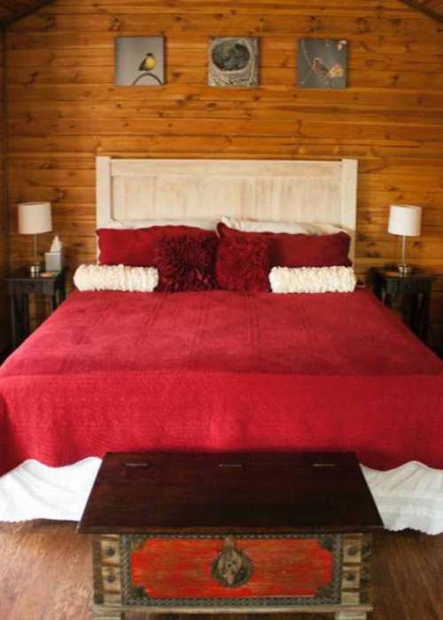 Cozy bedroom featuring a red bedspread, decorative pillows, wooden walls, and framed artwork above the bed.