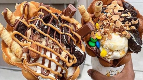 Two decadent ice cream cones topped with chocolate, cookies, nuts, and colorful candies, held in a hand.