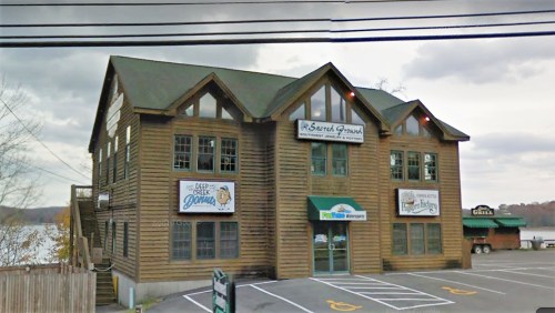 A large wooden building with multiple signs, located by a lake, featuring a restaurant and outdoor seating area.