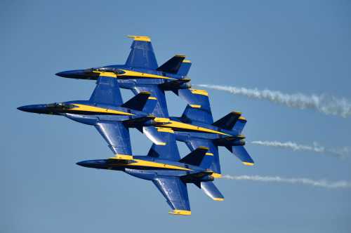 Four blue and yellow fighter jets flying in a tight formation, leaving trails of smoke against a clear blue sky.