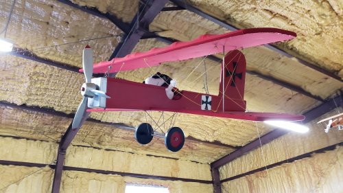 A red model airplane with a propeller hangs from the ceiling in a room with yellow insulation.
