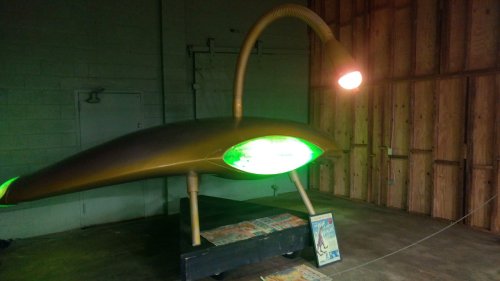 A futuristic, green-lit sculpture resembling a spaceship, displayed in a wooden interior space.