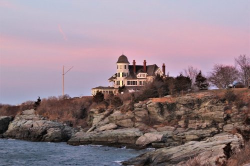 A large, elegant house sits atop rocky cliffs by the ocean, with a pastel sky at sunset in the background.