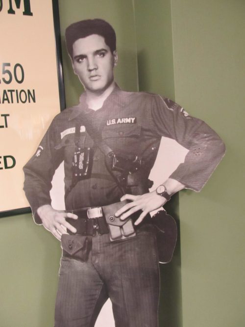 A life-sized cutout of Elvis Presley in a U.S. Army uniform, standing confidently with hands on hips.