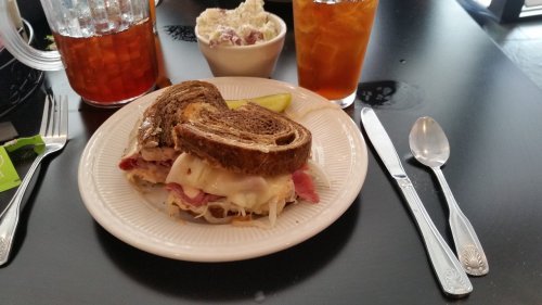 A plate with a sandwich on rye bread, a side of pickles, and a bowl of dessert, accompanied by iced tea.