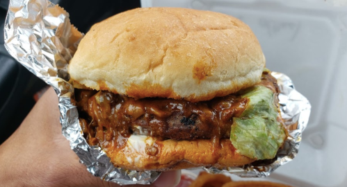 A hand holding a burger wrapped in foil, featuring a beef patty, lettuce, and a rich sauce on a soft bun.