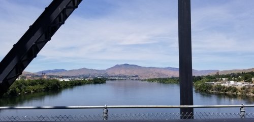 View of a calm river surrounded by greenery and mountains under a clear blue sky, framed by a bridge structure.