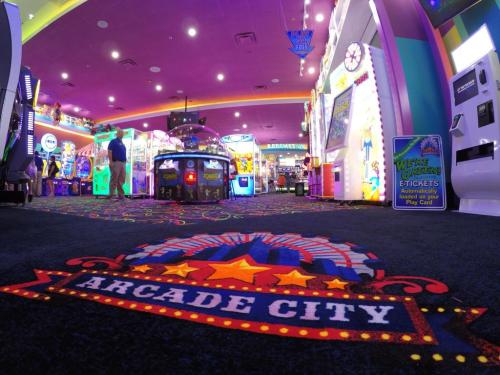 Colorful arcade interior with games, bright lights, and a carpet featuring "Arcade City" logo.