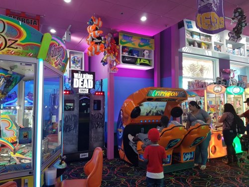 A colorful arcade filled with games, including a "Walking Dead" machine and a racing game, with people enjoying their time.
