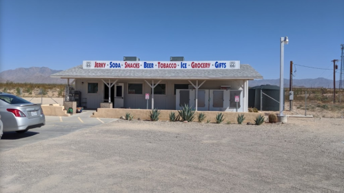 A small convenience store with a sign listing items for sale, set in a desert landscape with mountains in the background.