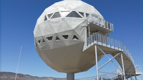 A large, geometric dome structure on a pedestal with a staircase, set against a clear blue sky and mountains in the background.