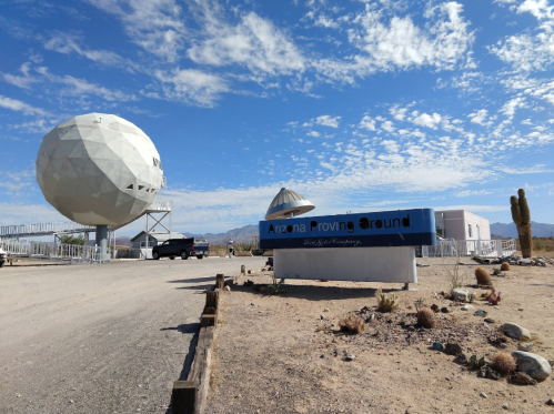 A large white dome structure and a sign reading "Arizona Proving Ground" against a blue sky with scattered clouds.