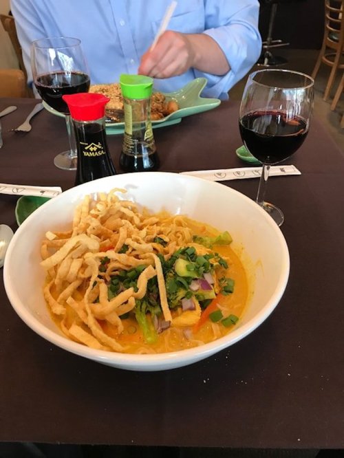 A bowl of curry noodles topped with crispy noodles, green onions, and two glasses of red wine on a table setting.