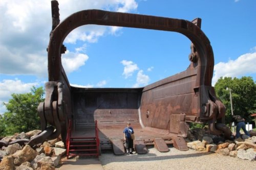 A large, rusted excavator bucket sculpture with a person standing inside, set against a blue sky and green trees.