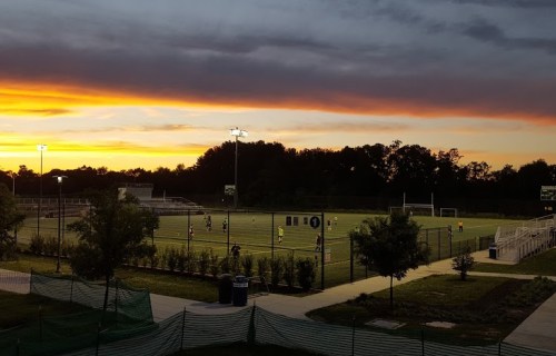 A soccer field at sunset, with players practicing under bright lights and a colorful sky in the background.